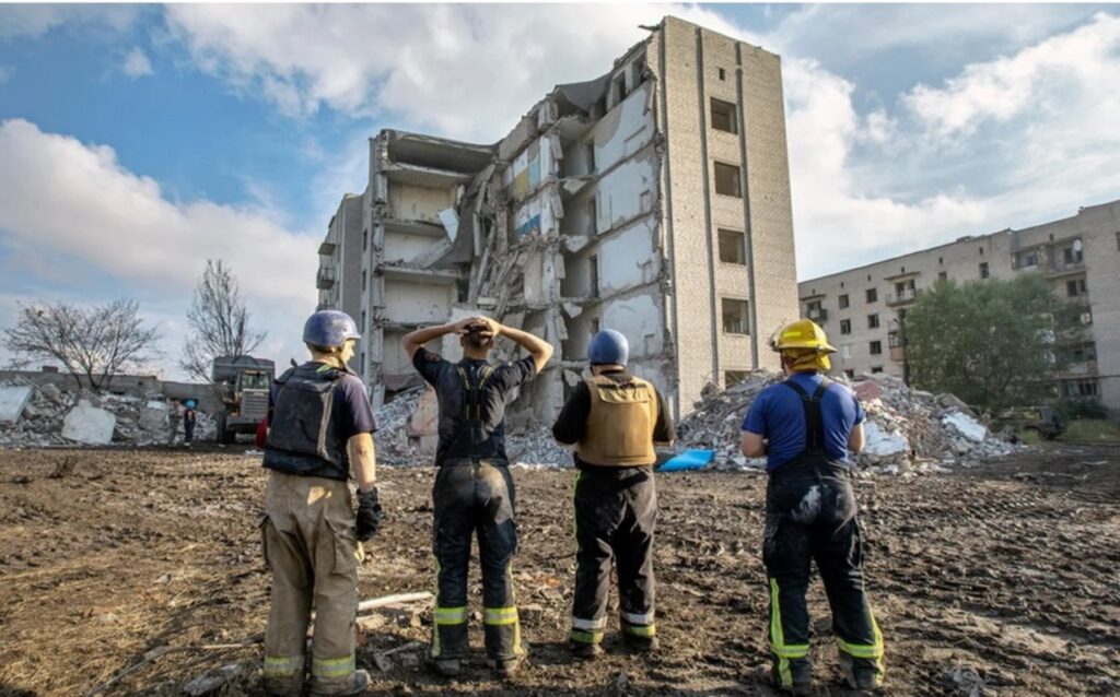 Ukrainos ugniagesiai susiėmę už galvų žiūri į sugriautą bendrabutį