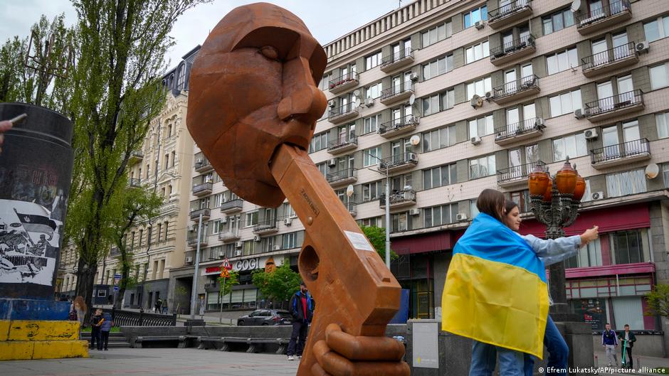 dvi merginos apsigaubusios Ukrainos vėliava pozuoja prie skulptūros, kurioje V. Putinas laiko įrėmęs ginklą sau į burną