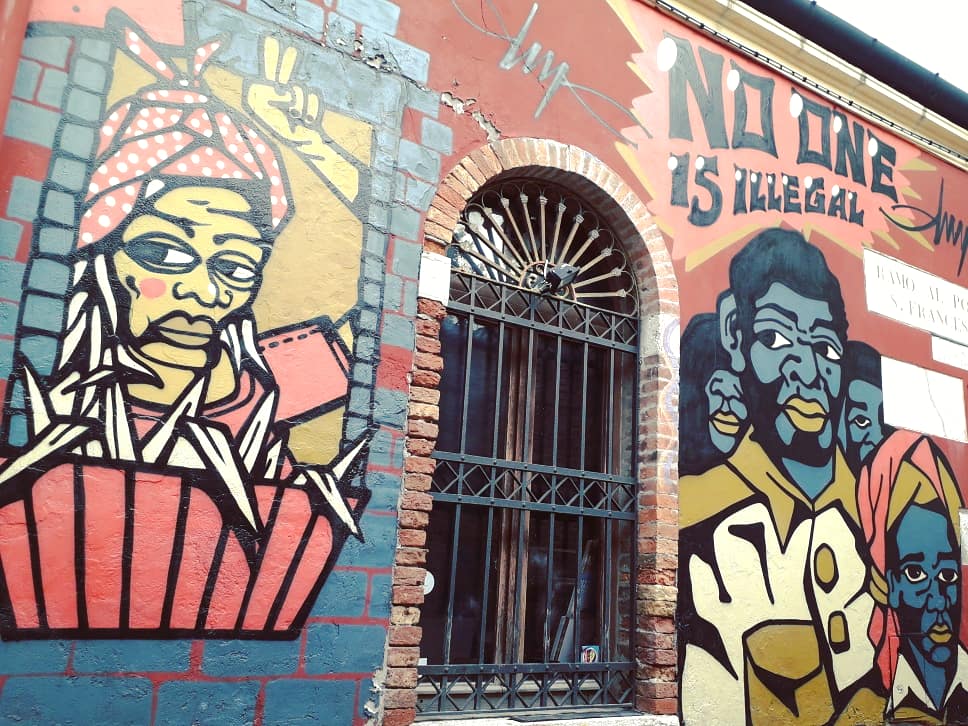 Grafitis, vaizduojantis du žmones, su užrašu „Niekas nėra nelegalus“