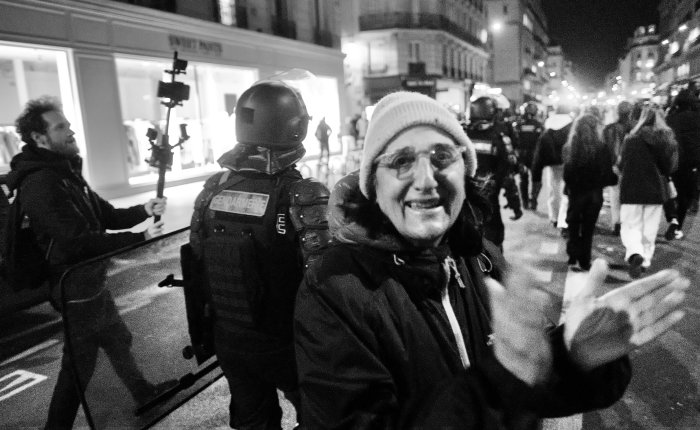 protestai Prancūzijoje: pagyvenusi moteris ploja, fone policija ir protestuotojai