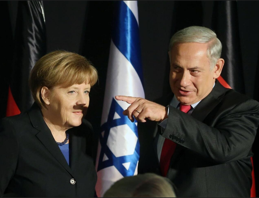Neytanyahu rodo pirštu, šalia stovi Angela Merkel, jai nuo piršto šešėlio pasimato Hitlerio ūsai
