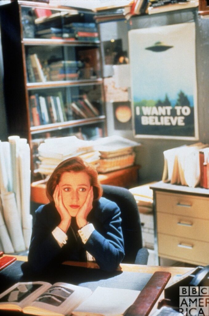 Dana Scully (Gillian Anderson) sėdi Foxo Mulderio kabinete pasirėmusi galvą rankomis, virš jos – plakatas „I want to believe“